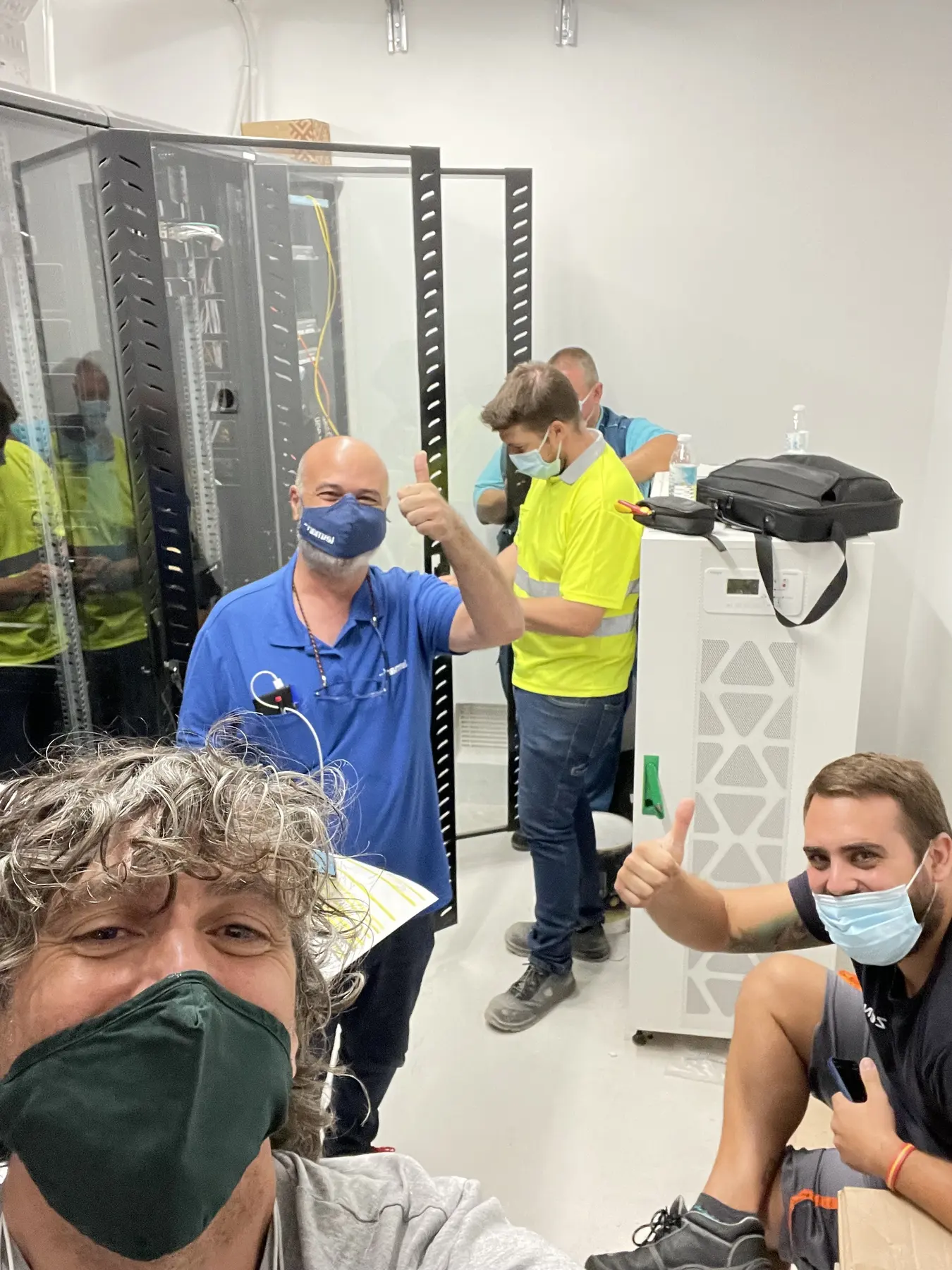Project team members giving a thumbs-up beside equipment racks in a data and utility room