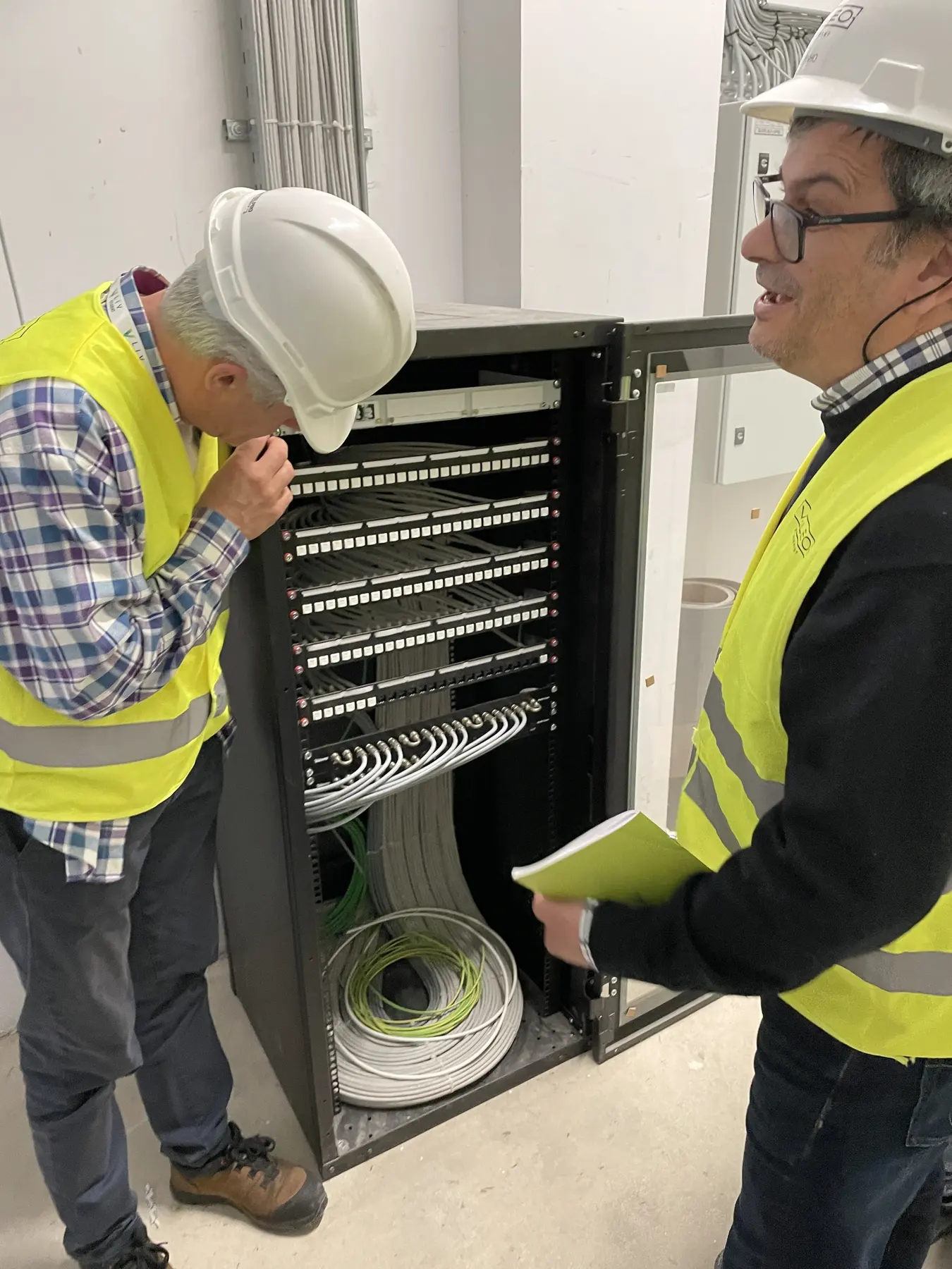 Two people in high-visibility vests and hard hats reviewing an open equipment rack in a utility room